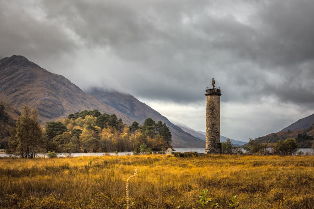 The Glenfinnan Monument standing tall against a dramatic cloudy Highland sky, with Loch Shiel and the mountains behind