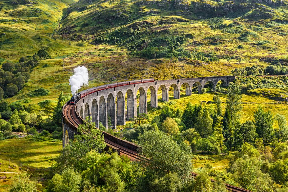 The Jacobite steam train crossing the Glenfinnan Viaduct, billowing white steam against lush green Highland hillsides